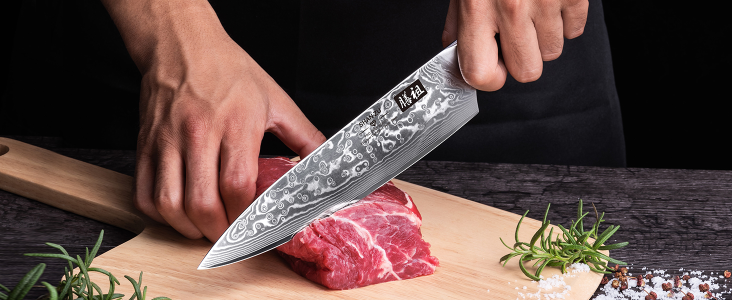 Les mains du chef utilisent un couteau en acier à motifs pour couper un morceau de viande rouge crue sur une surface foncée. Des brins de romarin sont visibles