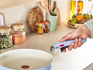 Versez le liquide à la main dans une poêle de couleur claire sur la cuisinière, avec des bocaux de rangement et des ustensiles de cuisine visibles en arrière-plan.