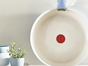 Poêle à frire en céramique blanche avec manche bleu clair accroché au mur. Petite plante herbacée en pot visible en arrière-plan. Logo circulaire rouge visible au centre du pan.